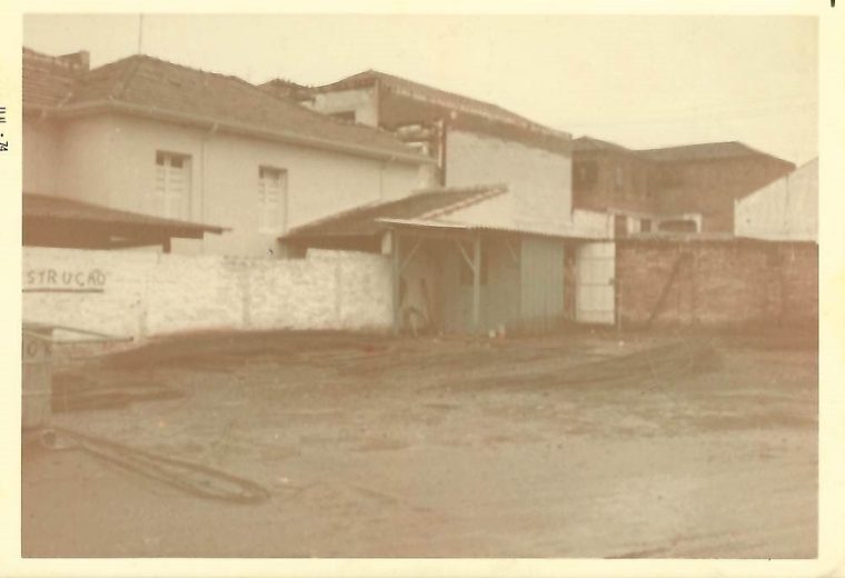 Terreno de terra batida com barracão de madeira e telha, muro de alvenaria ao fundo, em 1974